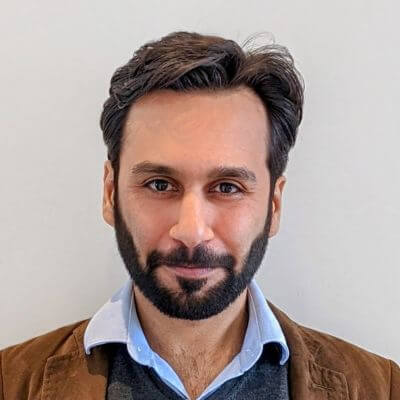 A headshot of Umar Naeem Ahmad, CEO of Abtrace. He has dark hair and a beard, and is wearing a light blue shirt under a dark sweater and brown jacket. He is looking at the camera with a slight smile.