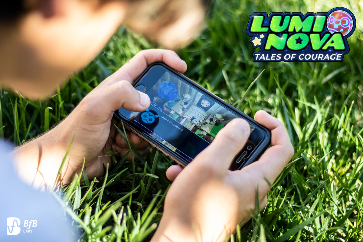 A close-up shot shows a person holding a smartphone horizontally, actively playing a game while sitting in green grass. The game screen displays colourful graphics, and the "Lumi Nova: Tales of Courage" logo is visible in the top right corner along with the "BfB Labs" logo in the bottom left.