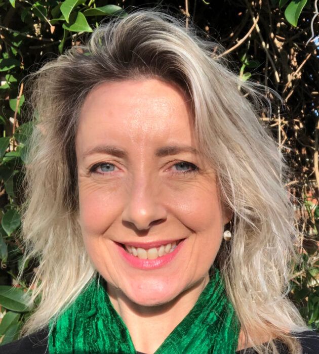 A close-up headshot of Louise Morpeth, the founder of Brain in Hand, shows her smiling directly at the camera. She has blonde hair and is wearing a vibrant green scarf. A leafy green bush is blurred in the background.