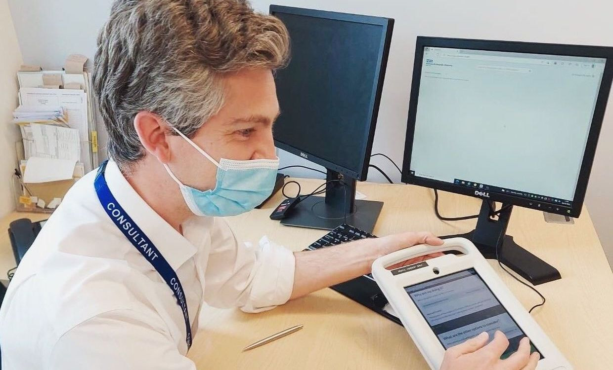 A consultant wearing a medical mask sits at a desk in front of two computer monitors. They are holding a white tablet device in their left hand while their right hand rests on a keyboard.