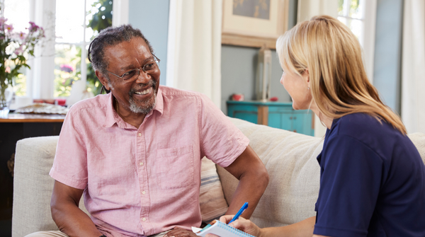 A man with greying hair and a beard, wearing glasses and a pink shirt, sits on a light-coloured sofa, smiling and looking at a blonde woman in a navy shirt who is sitting opposite him, writing on a notepad. They appear to be engaged in a conversation in a comfortable home setting.